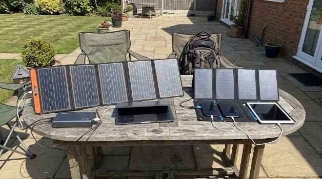 Flex Solar 60W and BigBlue 28W portable solar panels set up side-by-side on an outdoor wooden patio table, charging a laptop, power bank, smartphones, and a tablet under direct sunlight.
